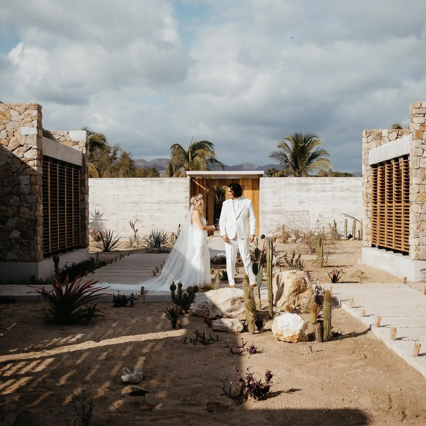 Bride and groom standing in a white stone courtyard with cacti and cloudy sky